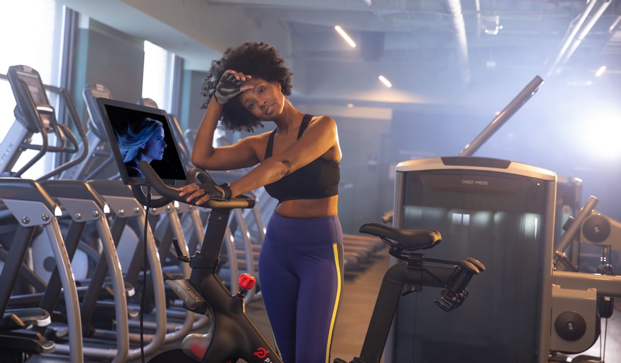 A woman in workout attire rests on an exercise bike in a gym, smiling and wiping her brow. The bike's screen shows a blue image. The gym is modern and well-lit.