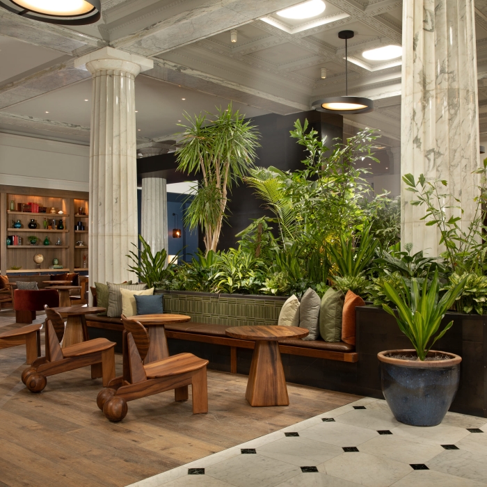 Spacious, elegant lobby with marble columns, lush indoor plants, wooden benches, and cozy seating. Warm, inviting atmosphere blending natural and modern elements.