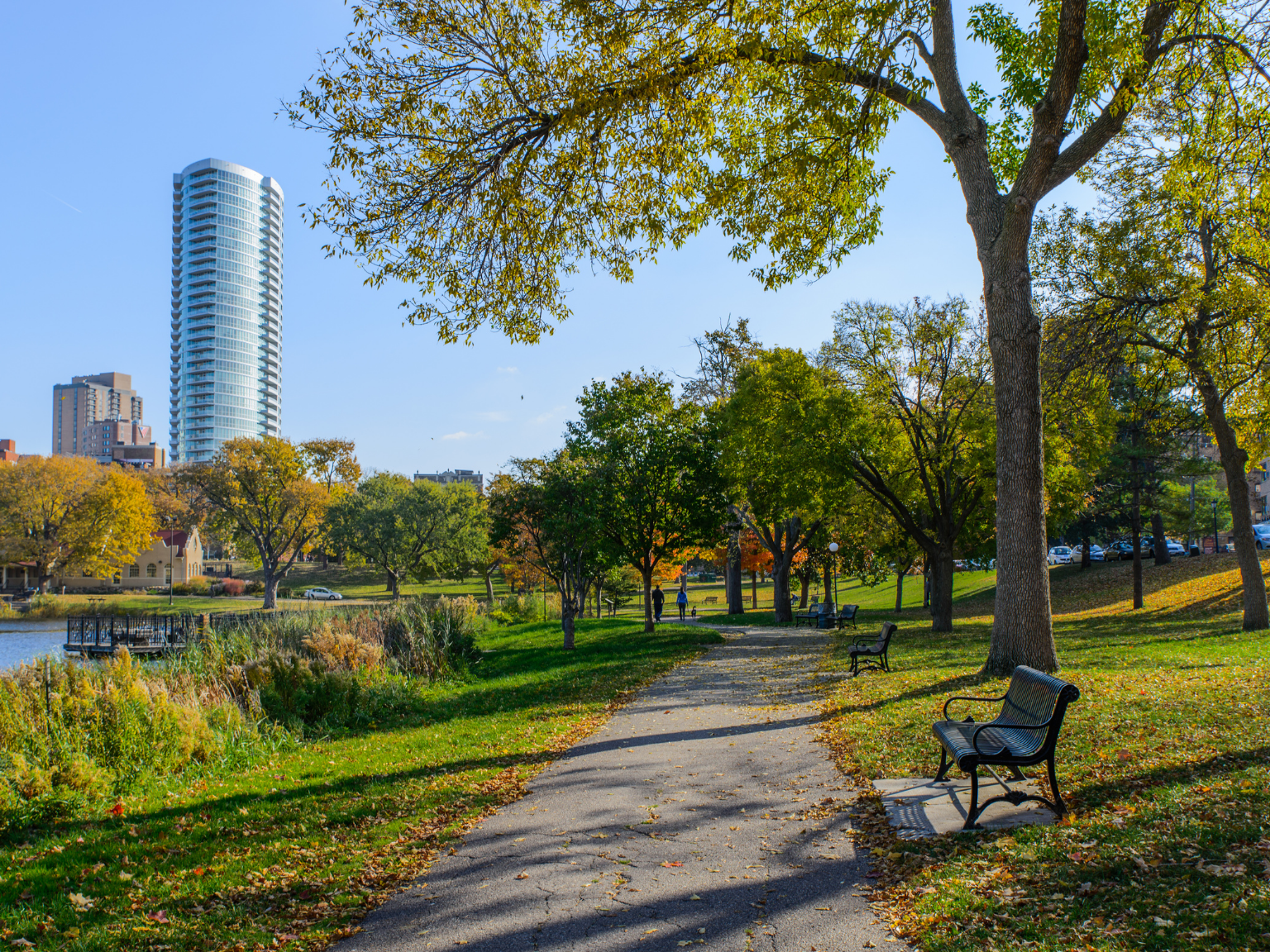 Park trail with a bench in the foreground and buildings in the background.