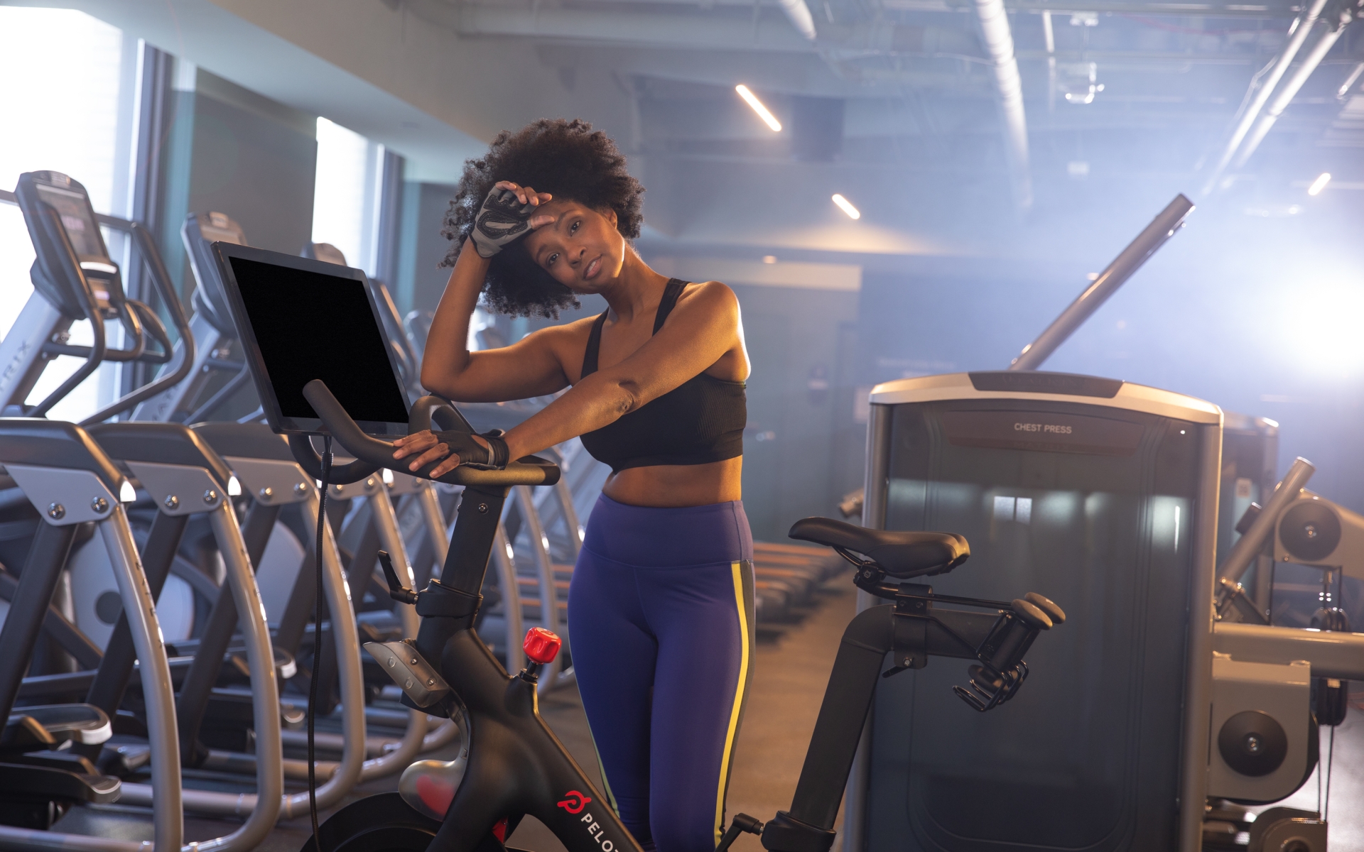 A woman in exercise clothes resting on a Peleton bike