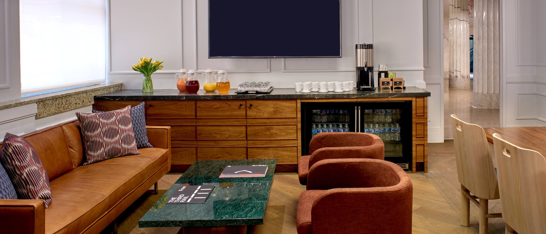 A cozy lounge with a brown leather sofa, colorful pillows, and a marble coffee table. A wooden cabinet holds drinks under a TV. Warm lighting and soft tones.