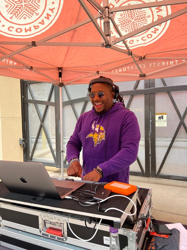 DJ wearing sunglasses and a purple hoodie, energetically mixes music on a laptop under a red canopy, creating a lively, upbeat atmosphere.