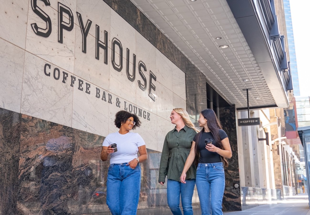 Three women walk along a downtown sidewalk outside Spyhouse Coffee Bar & Lounge, smiling and holding coffee cups, with the café’s large marble exterior sign visible above them.