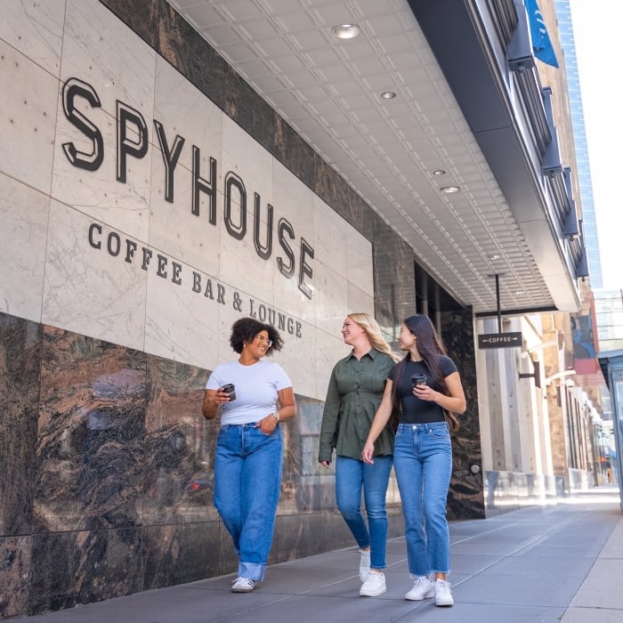 Three women walk along a downtown sidewalk outside Spyhouse Coffee Bar & Lounge, smiling and holding coffee cups, with the café’s large marble exterior sign visible above them.
