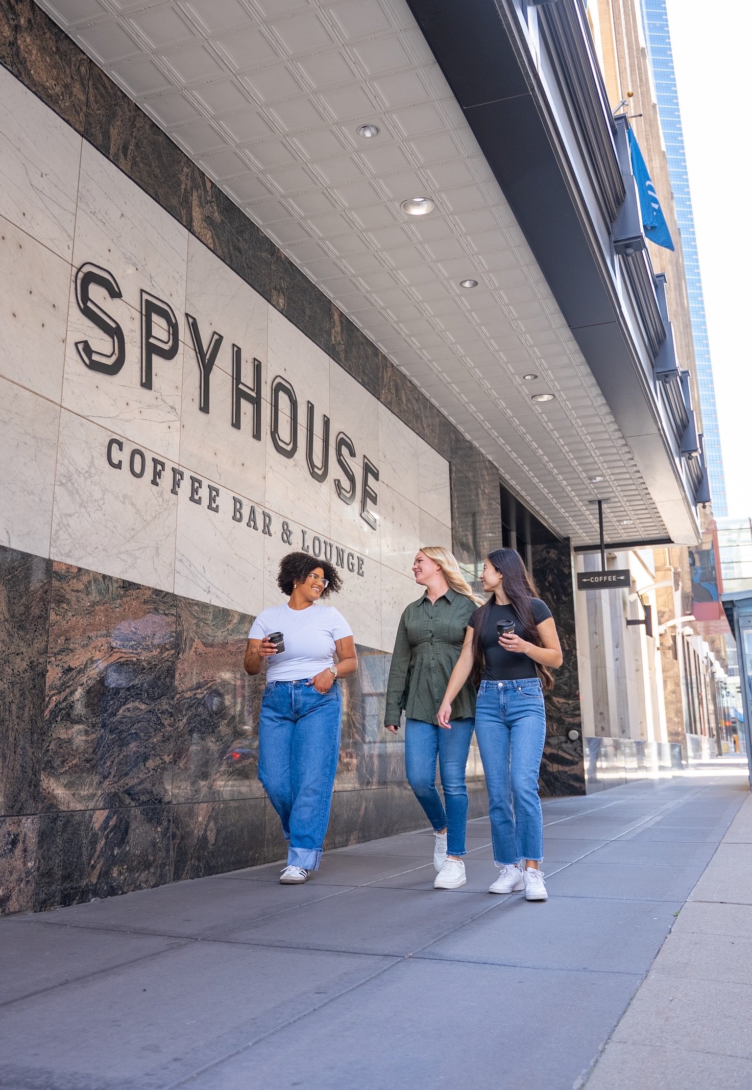 Three women walk along a downtown sidewalk outside Spyhouse Coffee Bar & Lounge, smiling and holding coffee cups, with the café’s large marble exterior sign visible above them.