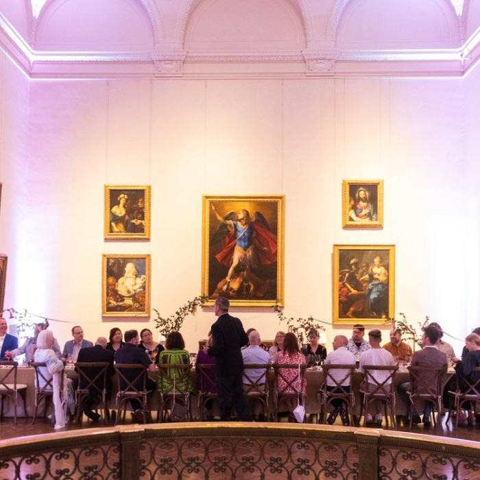 A large dining room features elegant high ceilings and classic paintings adorning the walls. Guests sit at a long table, suggesting a formal event.