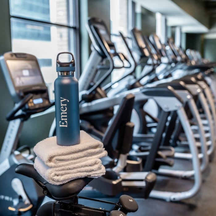 Emery branded water bottle sitting on stack of towels on top of Peloton bike with fitness center equipment in background