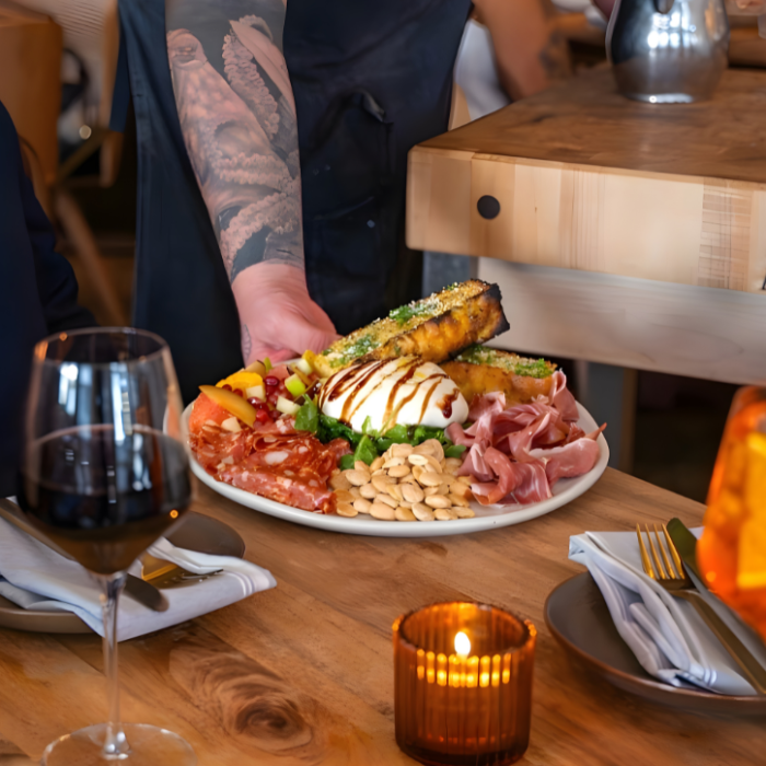 Hand of chef setting plate of fresh mozzarella, meats, nuts, and focaccia on to table with drinks