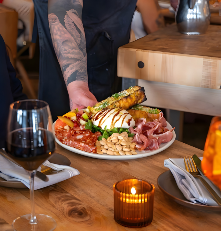 Hand of chef setting plate of fresh mozzarella, meats, nuts, and focaccia on to table with drinks